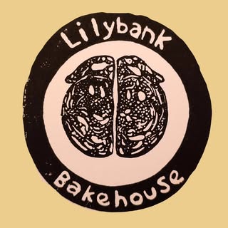 A circular black and white logo features the words Lilybank Bakehouse surrounding an illustration of a round, sliced loaf of bread with detailed texture—a nod to Montrose High Street and its vibrant revival. The background is light brown.