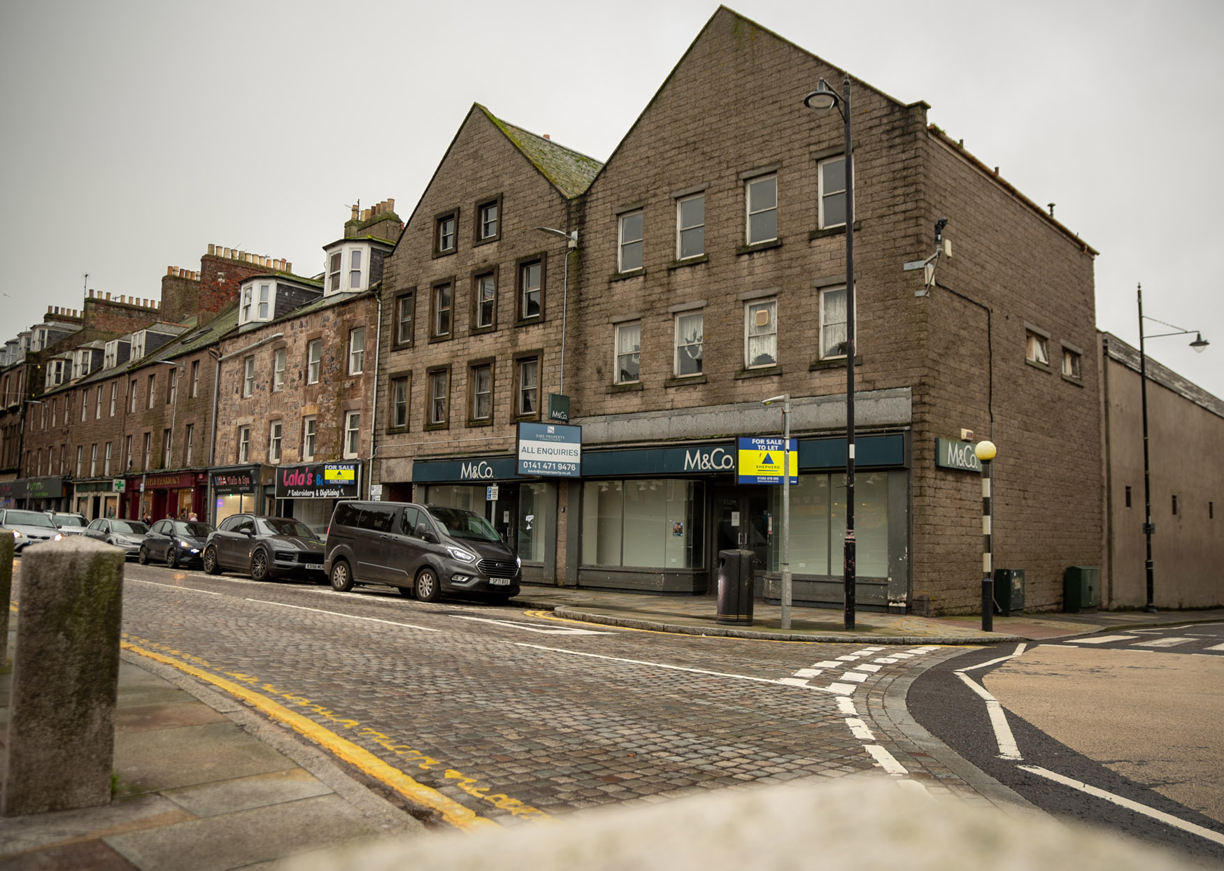 A quiet section of Montrose High Street, with stone buildings and cobbled paving, features shops like M&Co and Ladbrokes. A few cars are parked along the kerb beneath an overcast sky, capturing a classic Montrose revival charm.