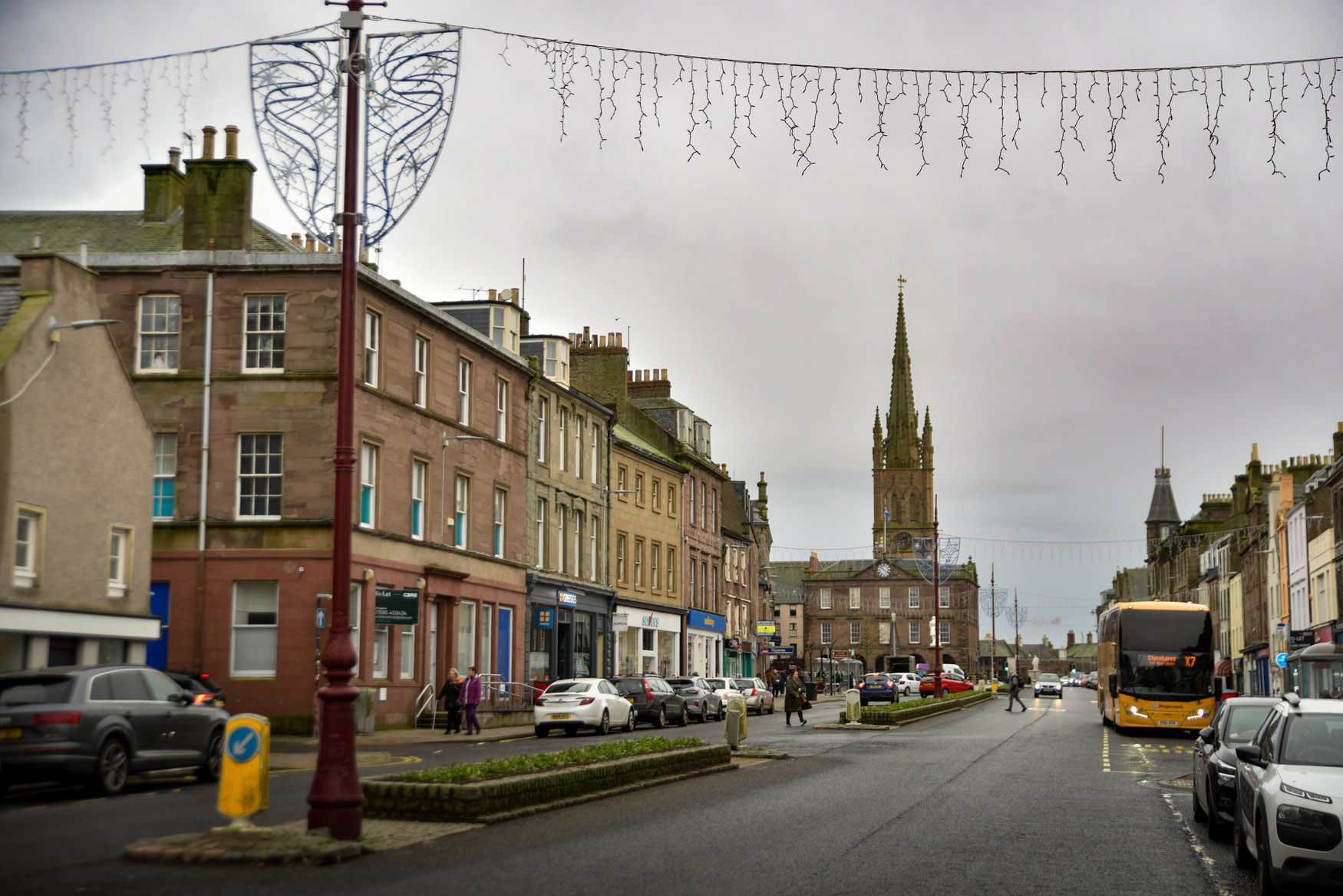 A street scene on Montrose High Street with parked cars, shops, and a church with a tall spire in the background. A bus is stopped on the right-hand side, and fairy lights hint at a Montrose Revival promo under the cloudy sky.