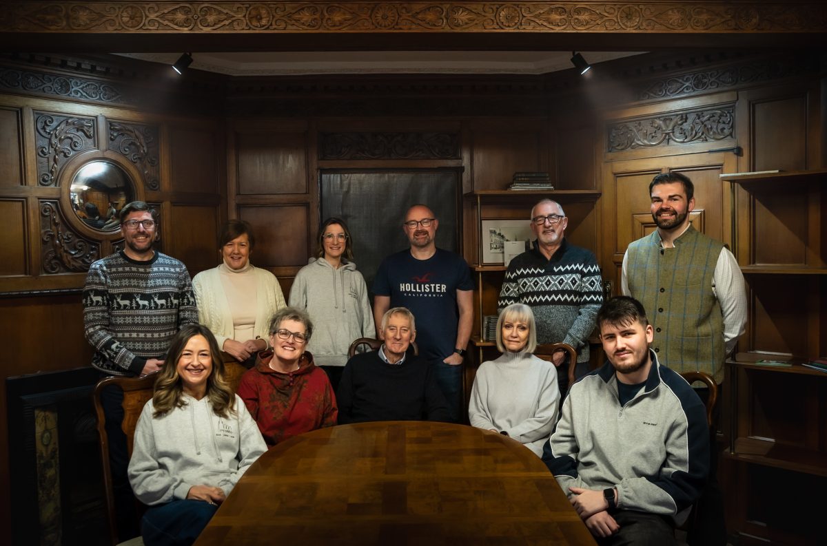 A group of eleven adults, some seated at a wooden table and others standing behind, pose for a promo photo in a room with ornate wooden panelling and shelves. The mood appears relaxed and casual, echoing the spirit of the Montrose revival.