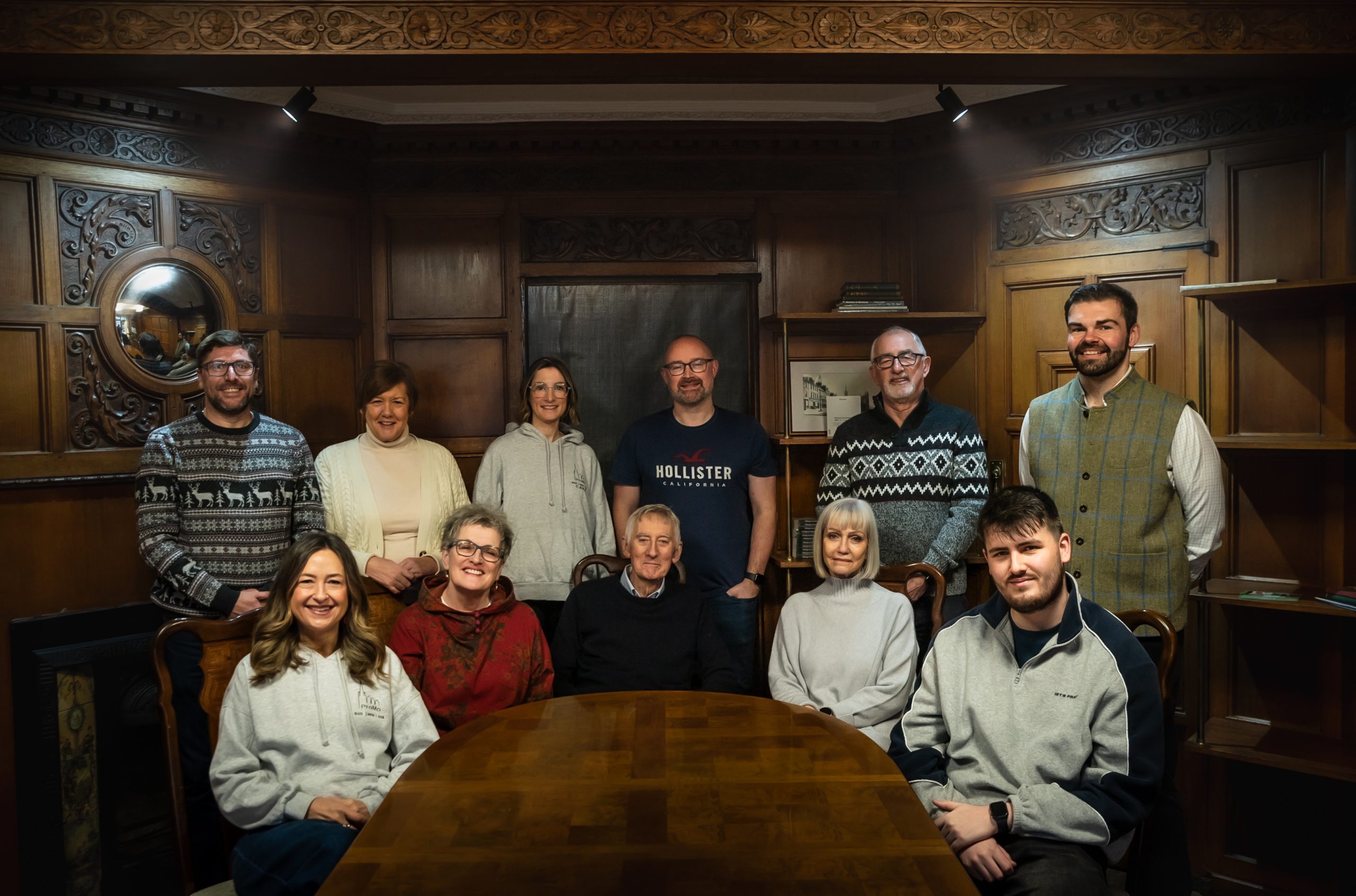 A group of eleven adults, some seated at a wooden table and others standing behind, pose for a promo photo in a room with ornate wooden panelling and shelves. The mood appears relaxed and casual, echoing the spirit of the Montrose revival.