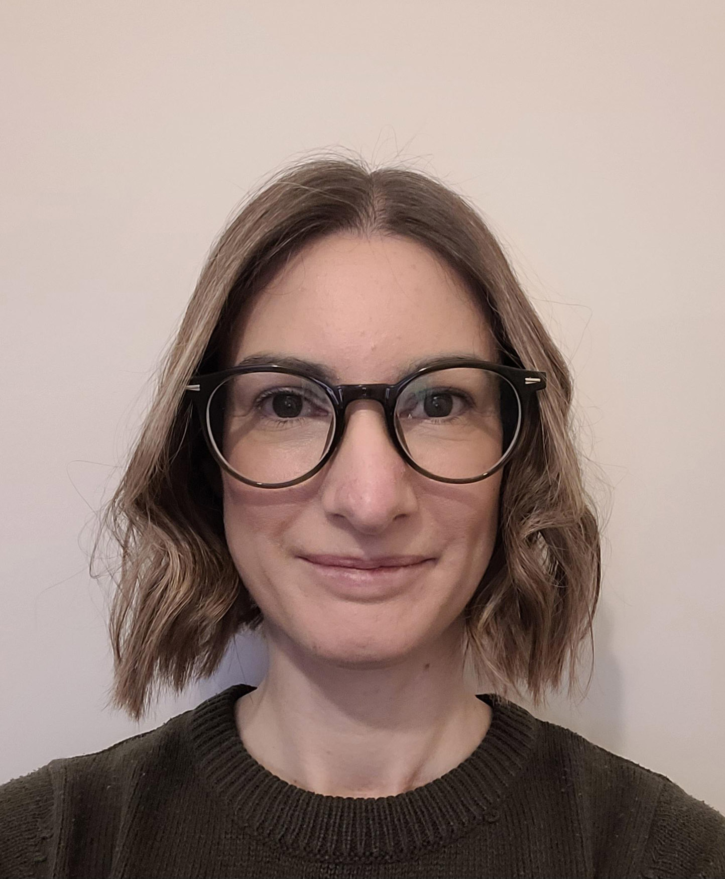 A person with shoulder-length light brown hair wearing large round glasses and a dark jumper is smiling slightly while facing the camera against a plain, light-coloured background.