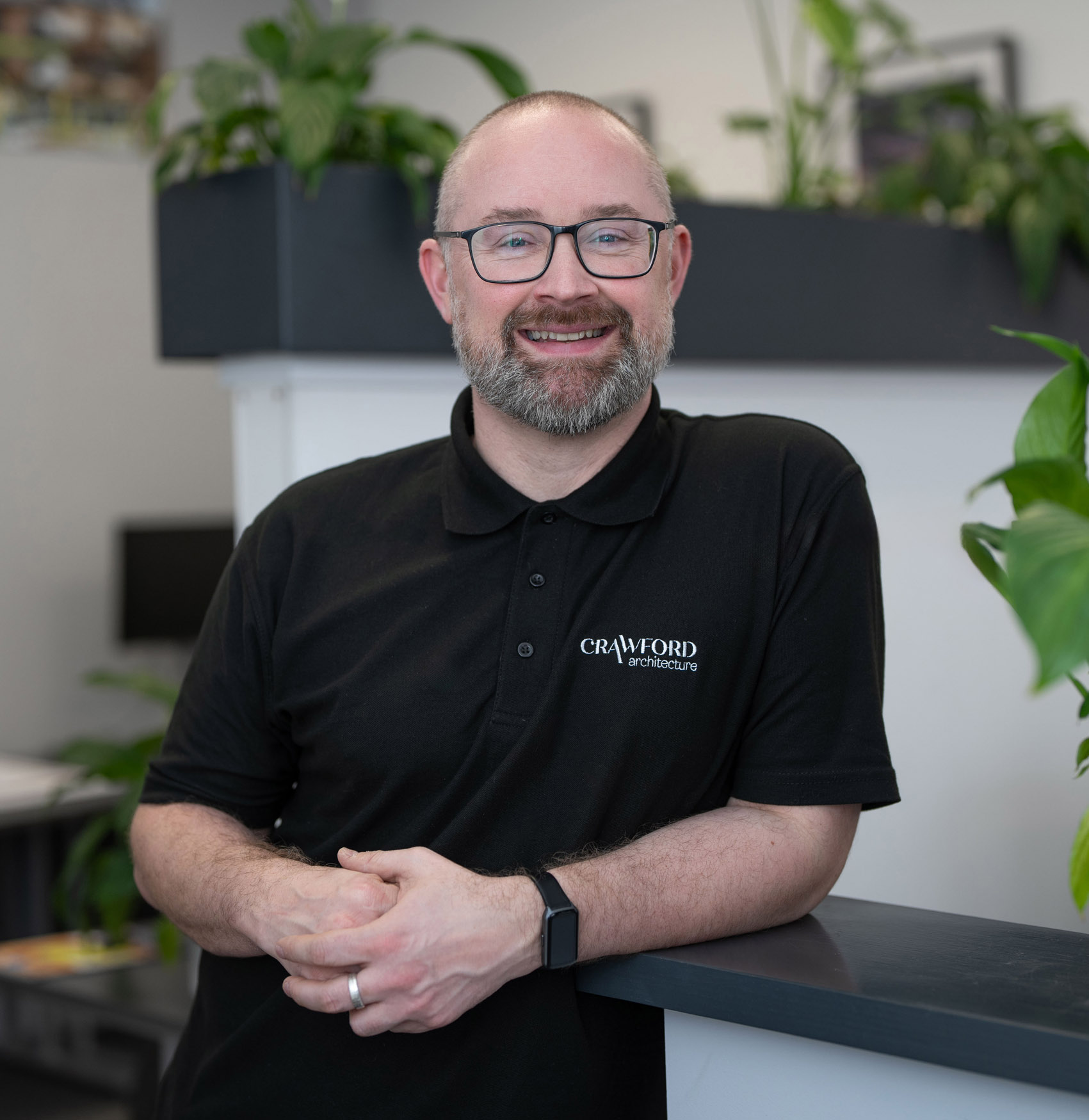 A man with glasses and a beard, wearing a black Crawford Architecture polo shirt and a smartwatch, stands indoors leaning on a counter, with green plants in the background.
