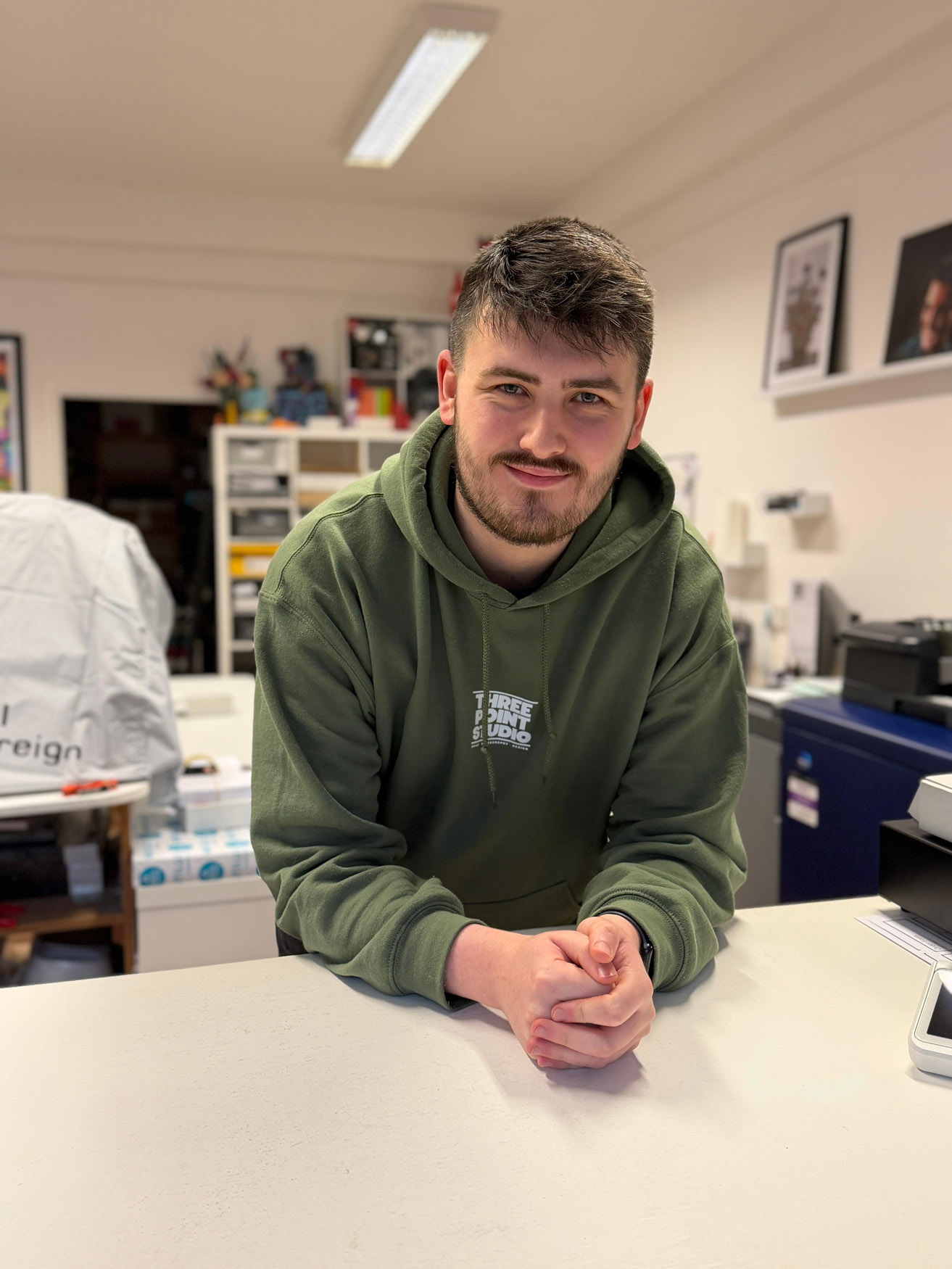 A man with short dark hair and a beard, wearing a green hoodie, stands behind a white counter in an office or shop, resting his forearms on the counter and smiling slightly at the camera. Shelves and office equipment are visible behind him.