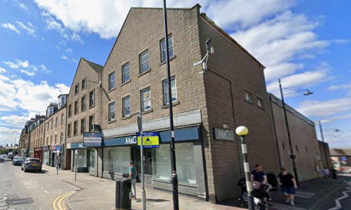 A street corner with a three-storey stone building featuring a shopfront labelled M&Co. Several people walk along the pavement, and a lamppost and street signs are visible. The sky is partly cloudy.