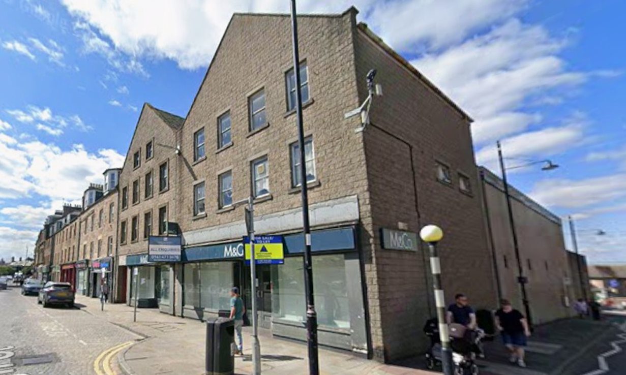 A street corner with a three-storey stone building featuring a shopfront labelled M&Co. Several people walk along the pavement, and a lamppost and street signs are visible. The sky is partly cloudy.