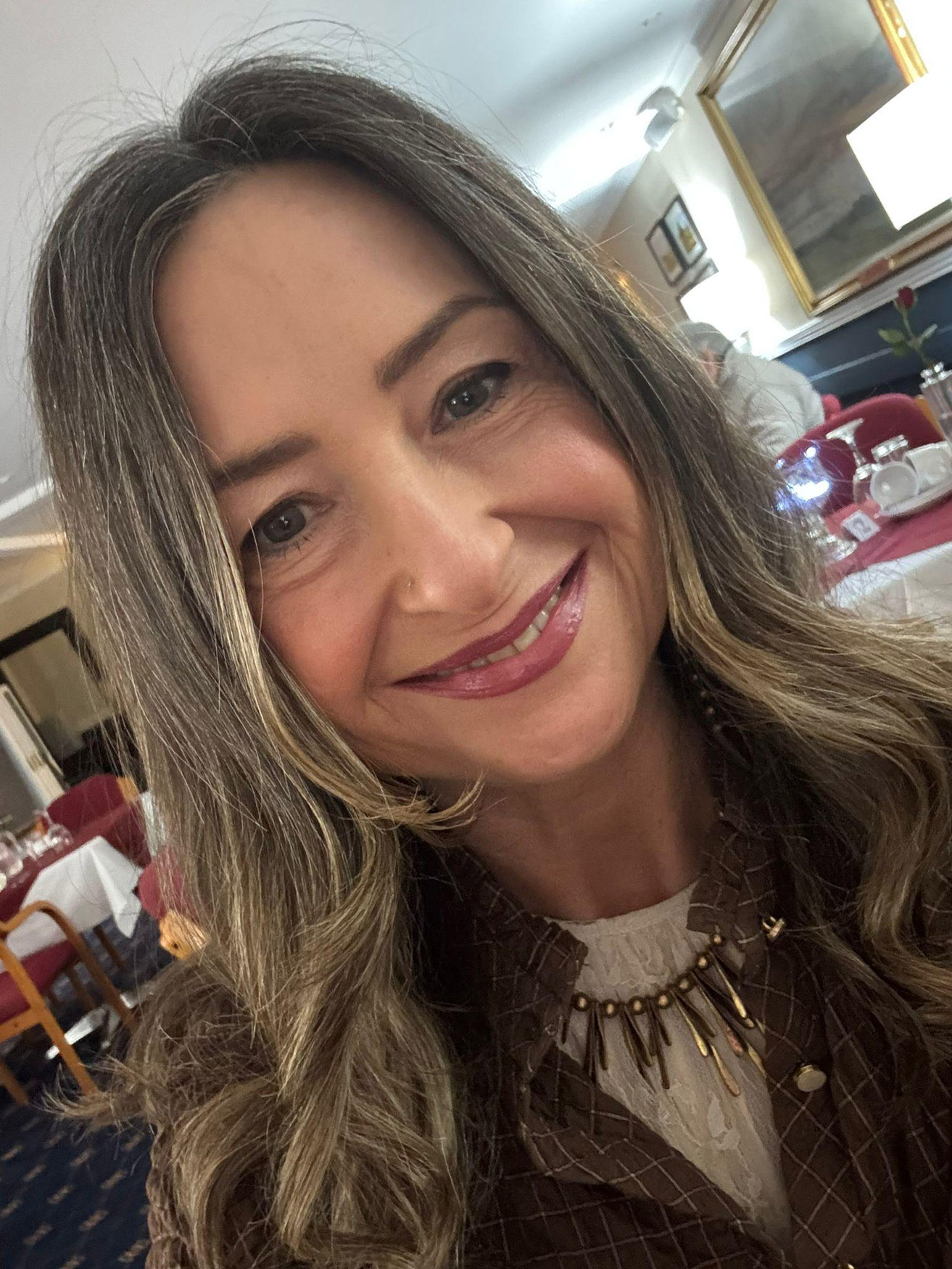A woman with long brown hair and a necklace smiles at the camera in a restaurant with red tablecloths, chairs, and framed artwork on the walls in the background.