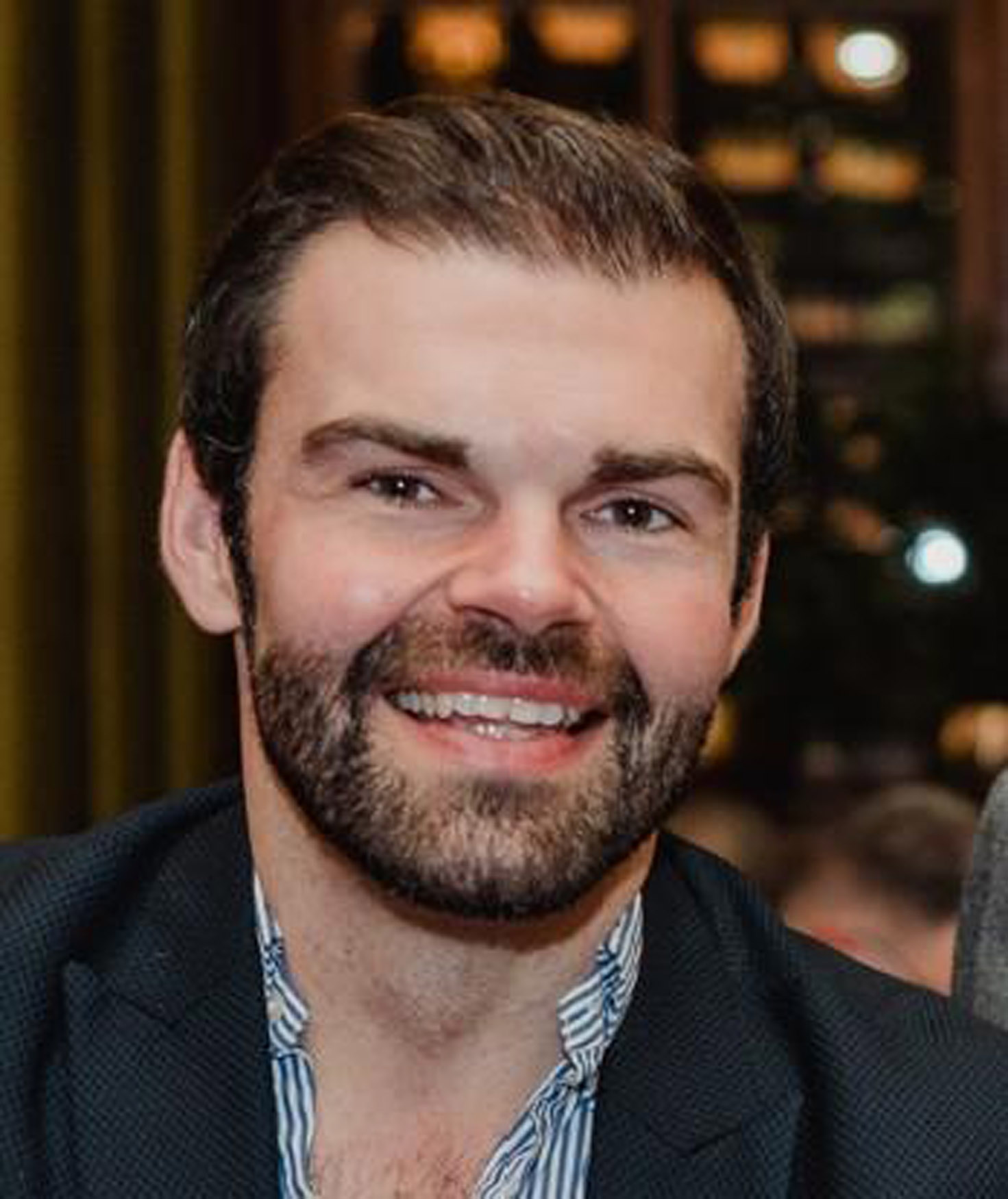 A man with dark hair and a beard is smiling. He is wearing a dark blazer over a blue and white striped shirt. The background is blurred, showing warm indoor lighting and what appear to be shelves or a cupboard.