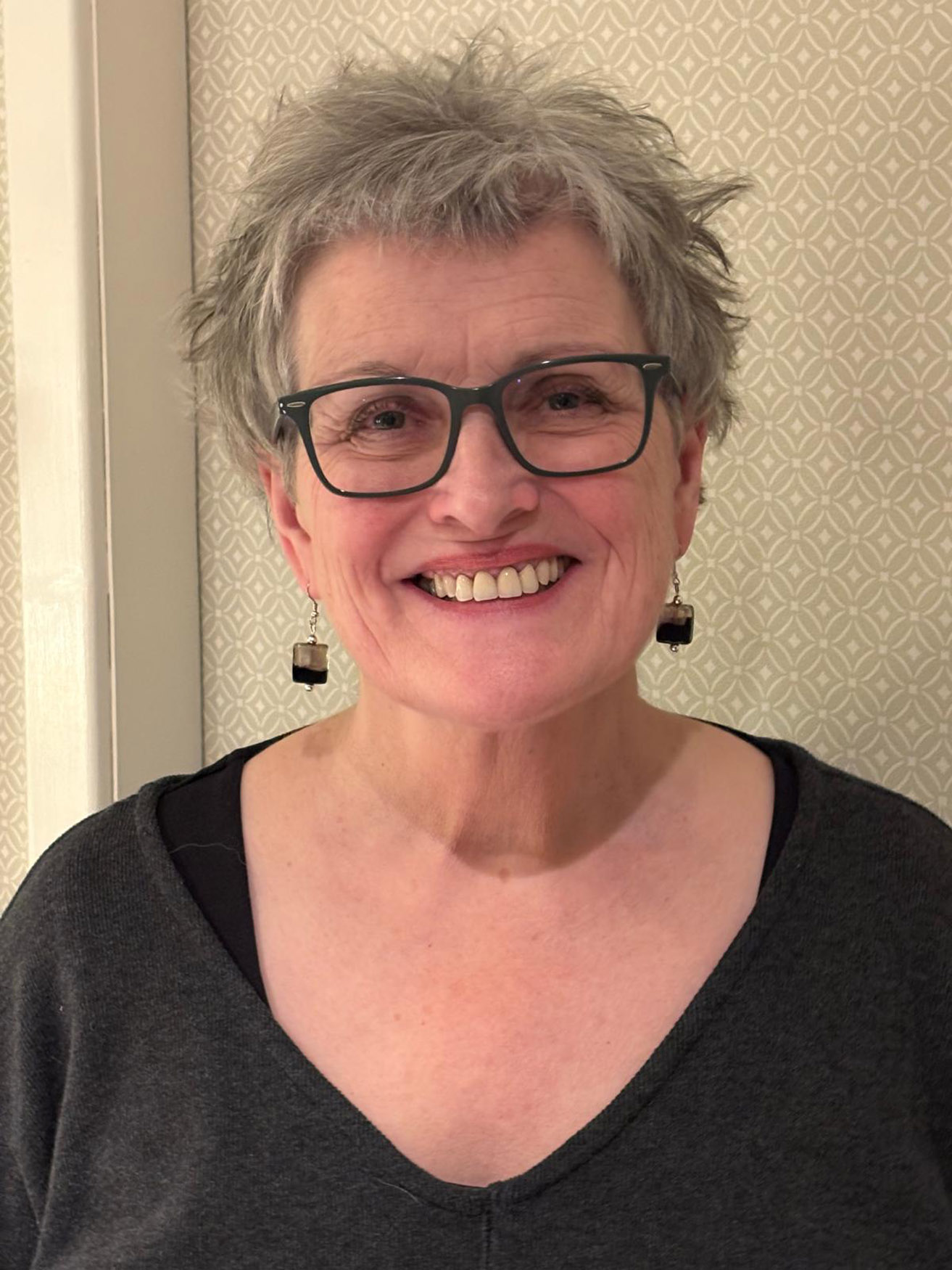 An older woman with short grey hair, glasses, and black earrings is smiling at the camera. She is wearing a dark top and standing in front of a patterned beige wall.