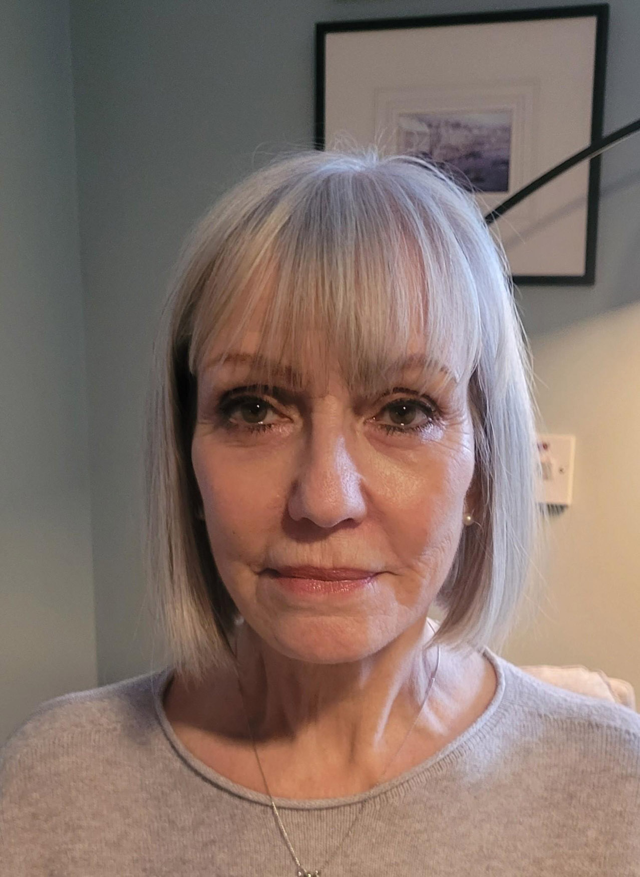 A woman with straight, shoulder-length grey hair and a fringe is facing the camera. She is wearing a light grey top and a necklace. There is a framed picture and a light switch on the wall behind her.