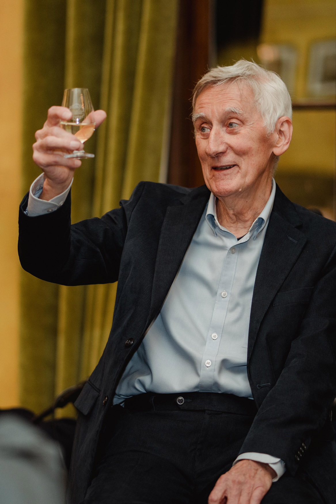 An older man with white hair wearing a dark suit and light blue shirt holds up a glass of white wine, appearing to make a toast. He is sitting in front of green curtains and smiling.