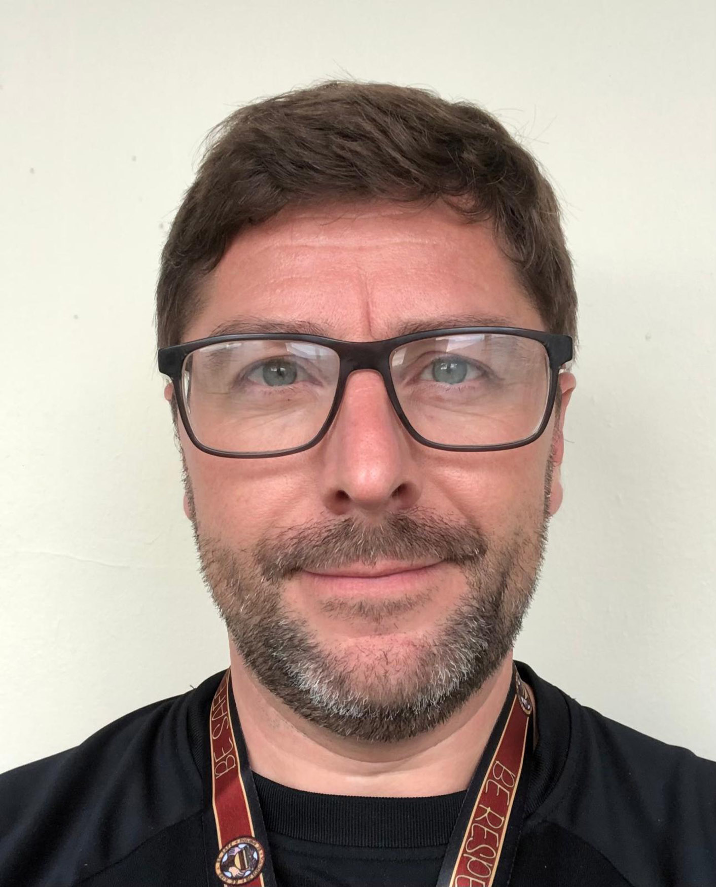 A man with short brown hair, a beard, and black-rimmed glasses is facing the camera. He is wearing a black shirt and a lanyard with text around his neck. The background is a plain off-white wall.