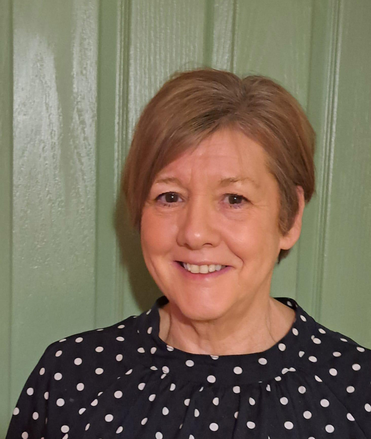 A woman with short light brown hair is smiling at the camera. She is wearing a black blouse with white polka dots and standing in front of a green panelled wall.